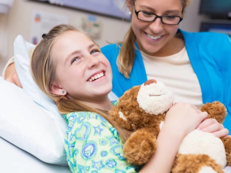 girl holding a teddy bear and smiling after her surgery with her mom nearby